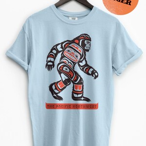 Puede incluir: Camiseta azul claro con un gr&aacute;fico de un Sasquatch estilizado en rojo, negro y blanco. El gr&aacute;fico est&aacute; rodeado del texto "The Pacific Northwest".