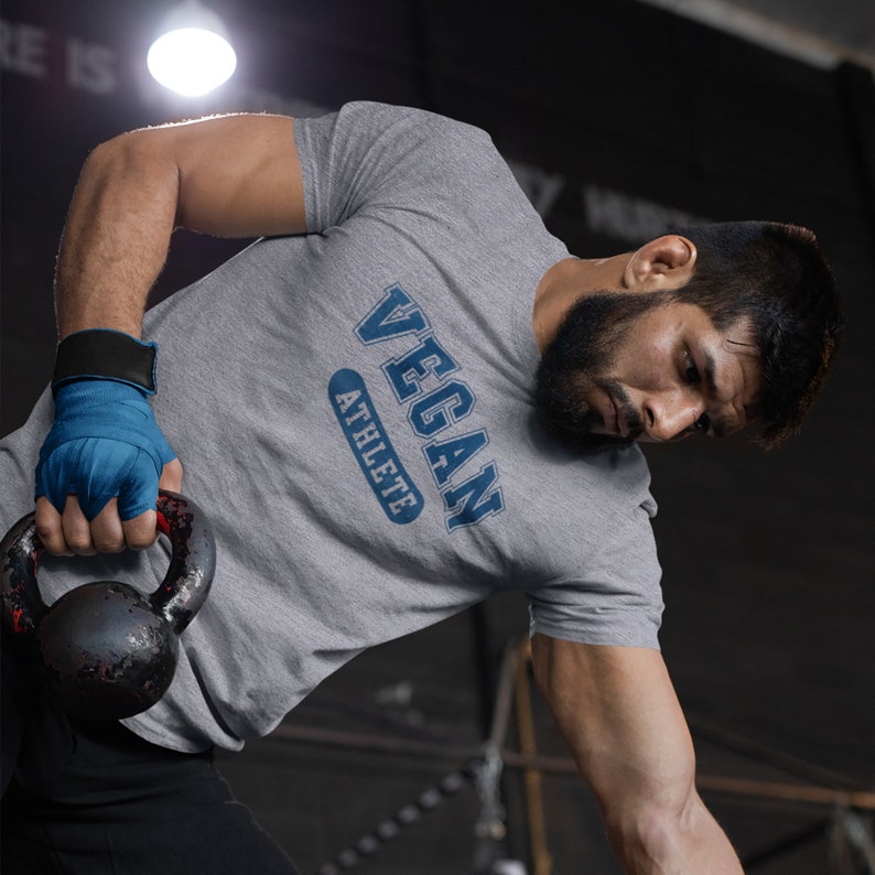 May include: A grey t-shirt with the text "VEGAN ATHLETE" in blue lettering. The person wearing the shirt is holding a black kettlebell.