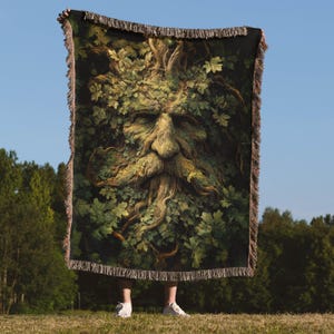 May include: A tapestry throw blanket featuring a detailed illustration of the Green Man, a face composed of leaves and branches. The blanket has a dark green background with a fringed edge, and is held up against a blue sky.
