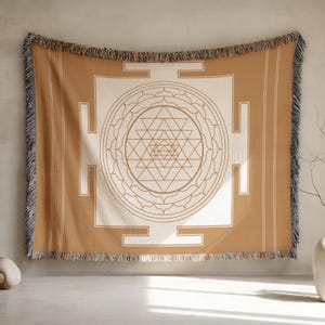 May include: A tan and cream-colored woven throw blanket with a geometric design. The blanket features a central geometric pattern with a series of triangles and circles. The edges are fringed.