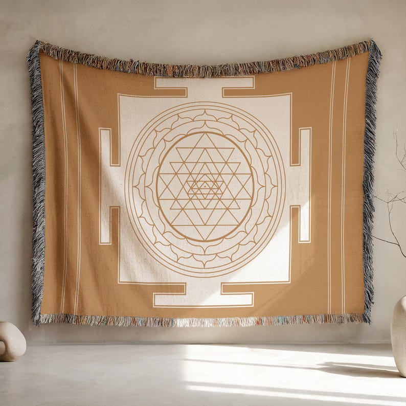 May include: A tan and cream-colored woven throw blanket with a geometric design. The blanket features a central geometric pattern with a series of triangles and circles. The edges are fringed.