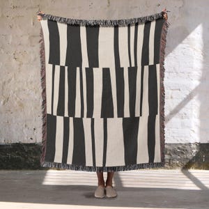 May include: A black and cream-colored woven throw blanket with a vertical stripe pattern. The blanket has a fringed edge in shades of brown and cream. The design features alternating thick and thin stripes.