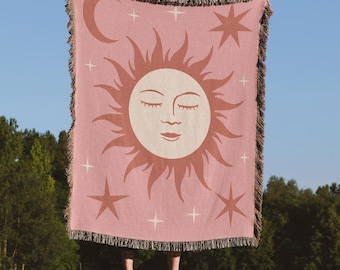 以下が含まれることがあります： ピンク色の織りブランケット。 太陽、月、星のデザイン。 太陽は目を閉じており、光線に囲まれています。 ブランケットはフリンジエッジです。 太陽と星は濃いピンク色です。