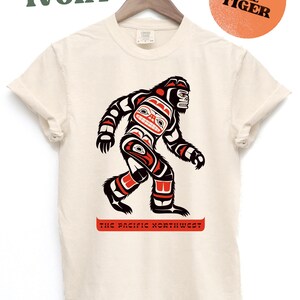 Puede incluir: Una camiseta de color crema con un gr&aacute;fico de un Sasquatch estilizado en rojo, negro y azul. El texto "The Pacific Northwest" est&aacute; impreso debajo del gr&aacute;fico. El texto "IVORY" est&aacute; impreso en verde en la parte superior de la imagen. El texto "BEN THE TIGER" est&aacute; impreso en negro sobre un c&iacute;rculo naranja quemado.