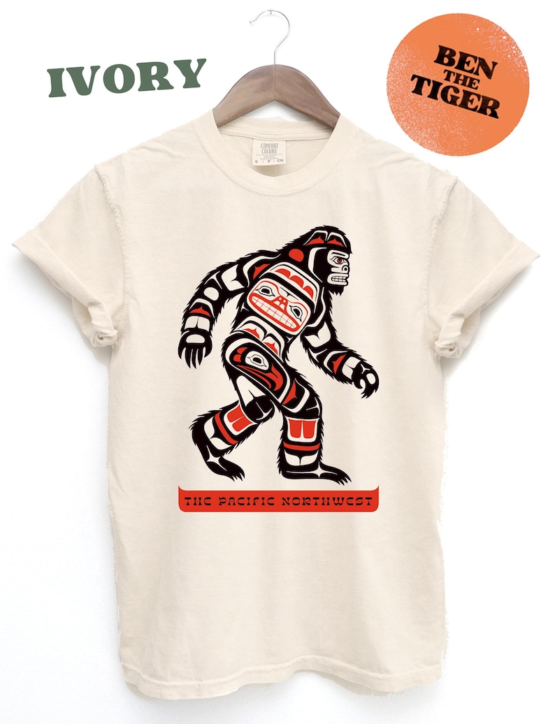 Puede incluir: Una camiseta de color crema con un gr&aacute;fico de un Sasquatch estilizado en rojo, negro y azul. El texto "The Pacific Northwest" est&aacute; impreso debajo del gr&aacute;fico. El texto "IVORY" est&aacute; impreso en verde en la parte superior de la imagen. El texto "BEN THE TIGER" est&aacute; impreso en negro sobre un c&iacute;rculo naranja quemado.