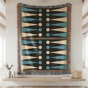 Op de afbeelding: Een grote, rechthoekige sprei met een geometrisch ontwerp. Het patroon heeft afwisselende driehoeken in lichtblauw en beige tegen een zwarte achtergrond, met beige en blauwe cirkels. De sprei heeft een bruine rand en franjes.