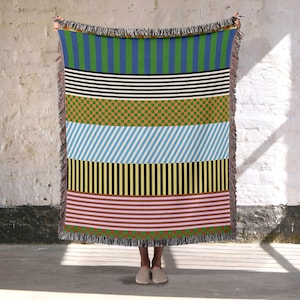 Peut inclure: Une couverture tissée colorée avec divers motifs géométriques. La couverture présente des rayures, des carreaux et des lignes diagonales dans des tons de bleu, vert, noir, blanc, jaune, marron et rose. Les bords sont finis avec une frange.