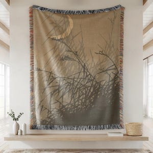 May include: A beige woven throw blanket with a nature-inspired design. The blanket features a silhouette of tall grass and a moon. The edges are fringed with a mix of colors. The blanket is displayed on a white wall.