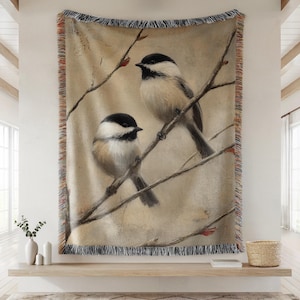 May include: A woven throw blanket featuring a painting of two chickadees perched on tree branches. The birds have black caps, white cheeks, and tan bodies. The blanket has a fringed edge with tan, orange, and black accents.