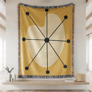 May include: Mustard yellow throw blanket with a mid-century modern design. The blanket features a large off-white shape with black lines radiating from a central point, and black dots. The edges are fringed.