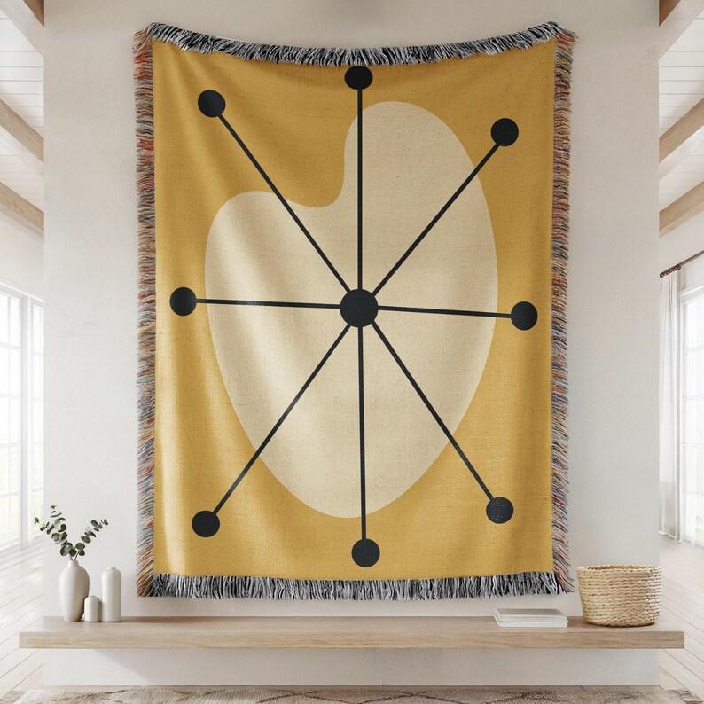 May include: Mustard yellow throw blanket with a mid-century modern design. The blanket features a large off-white shape with black lines radiating from a central point, and black dots. The edges are fringed.