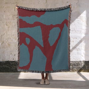 May include: A teal and burgundy throw blanket with a tree-like pattern. The blanket has a fringed edge and is held up against a white brick wall. The design features a teal background with burgundy abstract shapes.