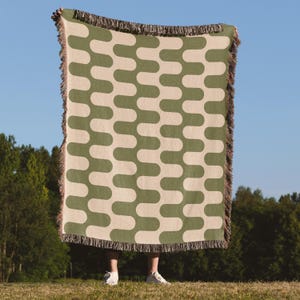 May include: A woven blanket with a repeating pattern of olive green and cream-colored curved shapes. The blanket has a fringed edge and is held up against a blue sky background. The blanket is displayed outdoors on a sunny day.
