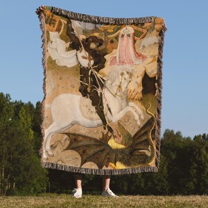 May include: A woven tapestry blanket featuring a medieval painting of a knight on horseback slaying a dragon. The blanket has a beige background with a detailed illustration, fringed edges, and is held up against a blue sky.