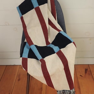 May include: A woven throw blanket draped over a chair. The blanket features a cream-colored background with bold stripes of burgundy, black, and light blue. The blanket has a fringed edge and is made of a soft, textured material.