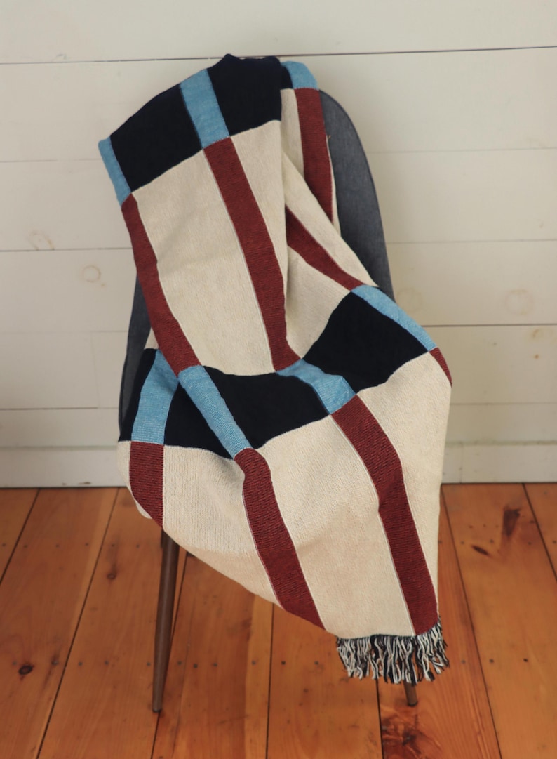 May include: A woven throw blanket draped over a chair. The blanket features a cream-colored background with bold stripes of burgundy, black, and light blue. The blanket has a fringed edge and is made of a soft, textured material.
