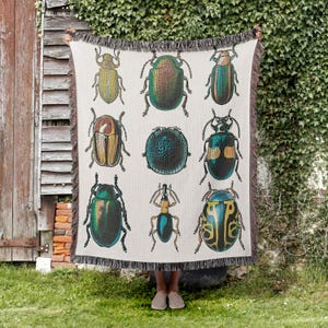 May include: Beige throw blanket featuring a repeating pattern of colorful beetle illustrations. The insects are depicted in shades of green, gold, and blue, with intricate details. The blanket has a fringed edge and is held up against a wooden door and ivy.