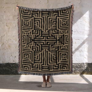 Könnte beinhalten: Eine beige gewebte Wurfadecke mit einem schwarzen geometrischen Muster. Das Design zeigt eine Reihe von miteinander verbundenen Linien und Formen, die ein labyrinthartiges Aussehen erzeugen. Die Decke hat einen Fransenrand und wird vor einer weißen Wand gehalten.
