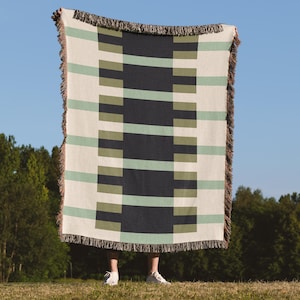 May include: A rectangular throw blanket with a geometric pattern. The blanket features alternating stripes of cream, navy, olive green, and light green. The edges are finished with a fringed trim. The blanket is held up against a blue sky background.