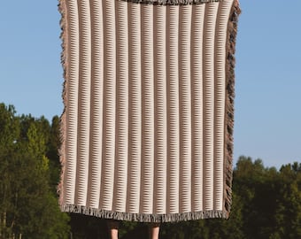 Könnte beinhalten: Eine beige gewebte Wurkdecke mit einem geometrischen Muster aus vertikalen Linien und Zickzack. Die Decke hat einen Fransenrand in Braun- und Beigetönen. Die Decke wird vor einem blauen Himmelshintergrund gehalten.