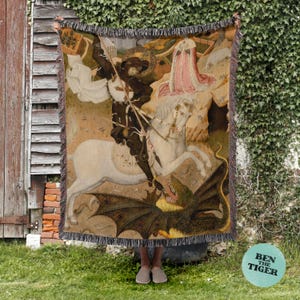 May include: Woven tapestry throw blanket featuring a medieval painting of a knight on horseback slaying a dragon. The artwork depicts a knight in dark armor, a white horse, and a dragon with large wings. The blanket has a fringed edge and is displayed outdoors.