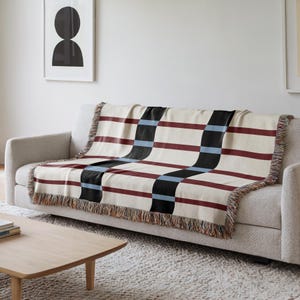 May include: A cream-colored throw blanket with a geometric pattern of burgundy, black, and light blue stripes. The blanket is draped over a gray sofa and has fringed edges. The design is modern and suitable for home decor.