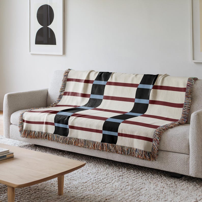 May include: A cream-colored throw blanket with a geometric pattern of burgundy, black, and light blue stripes. The blanket is draped over a gray sofa and has fringed edges. The design is modern and suitable for home decor.