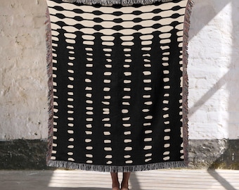 May include: A black and cream-colored woven throw blanket with a geometric pattern of horizontal ovals and vertical dashes. The blanket has a fringed edge and is held up against a white brick wall.
