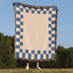 May include: A beige and blue checkered throw blanket with fringed edges is held up against a blue sky. The blanket has a large beige center with a border of blue squares. The fringe is a mix of beige and blue.