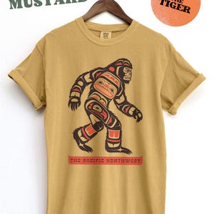Puede incluir: Camiseta amarilla mostaza con un gr&aacute;fico de un Sasquatch estilizado al estilo del arte ind&iacute;gena de la costa noroeste. El gr&aacute;fico incluye el texto "The Pacific Northwest".