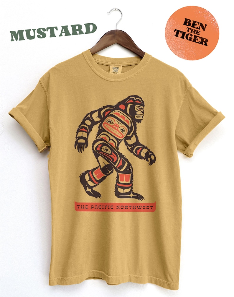 Puede incluir: Camiseta amarilla mostaza con un gr&aacute;fico de un Sasquatch estilizado al estilo del arte ind&iacute;gena de la costa noroeste. El gr&aacute;fico incluye el texto "The Pacific Northwest".
