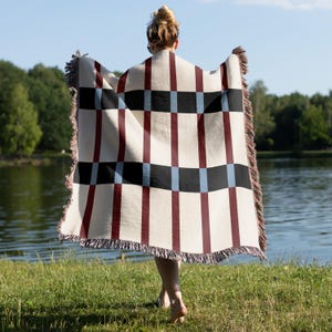 May include: A cream-colored woven blanket with a geometric pattern of burgundy, black, and light blue rectangles and stripes. The blanket has fringed edges and is held by a person standing near a lake.