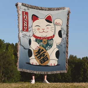 May include: A woven throw blanket featuring a Japanese Maneki Neko cat design. The cat is white with black spots, red accents, and a gold coin. The word "LUCKY" is in a red rectangle. The blanket has a fringed edge.