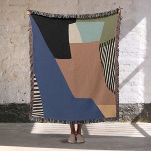 May include: A woven throw blanket with an abstract geometric design. The blanket features a colour block pattern with shades of blue, black, brown, and green, accented by black and white stripes. The edges are finished with fringe.