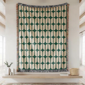 May include: A large, rectangular woven throw blanket with a repeating pattern of green and cream-coloured ovals. The blanket is fringed on the edges and hangs on a white wall. The throw blanket is a decorative home textile.