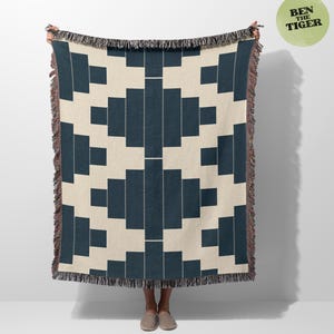May include: A woven throw blanket with a geometric pattern in navy blue and cream. The design features rectangular shapes and vertical lines of small white dots. The blanket has a fringed edge and is held up by a person.