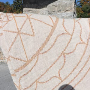 May include: Cream-colored woven textile with a geometric pattern in shades of tan and light brown. The design features intersecting lines and curved shapes, creating a modern, abstract look. The fabric appears to be thick and textured, suitable for a rug or blanket.