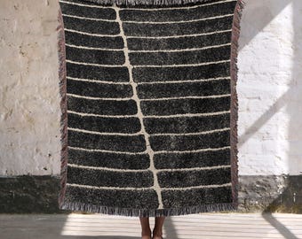 May include: A black and white woven throw blanket with a repeating horizontal stripe pattern and a central vertical design. The blanket has a fringed edge with a hint of pink. The blanket is held up in front of a white wall.