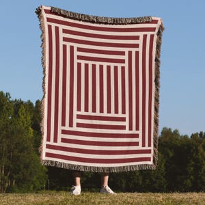 May include: A red and white striped throw blanket with a geometric design. The blanket has a fringe trim and is held up against a blue sky background. The blanket's pattern consists of vertical and horizontal stripes.