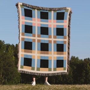 May include: A woven throw blanket featuring a grid pattern of large black squares separated by cream-coloured bands. The design includes accent blocks in light blue, soft pink, tan, and olive green, finished with a fringed edge on all four sides.