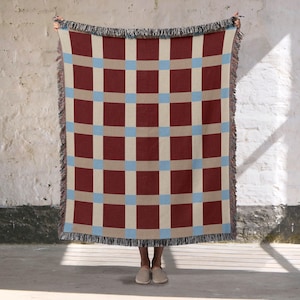 May include: A woven throw blanket featuring a grid pattern of large dark red rectangles separated by light beige bands and small light blue squares at the intersections. The blanket has a decorative fringed edge along the top and bottom sides.