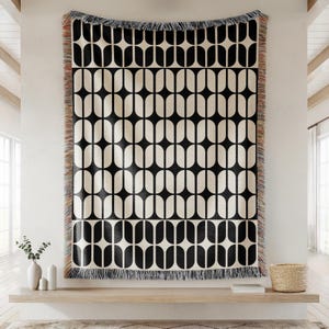 May include: A large, rectangular black and cream-colored throw blanket with a repeating geometric pattern of elongated ovals. The blanket is fringed along the edges and hangs on a white wall. A wooden shelf with decor is below.
