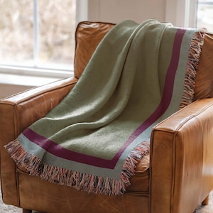 May include: A sage green woven throw blanket featuring a deep burgundy border stripe and multi-colored fringed edges. The textile has a textured appearance with subtle flecks throughout the fabric, draped over a brown leather armchair.