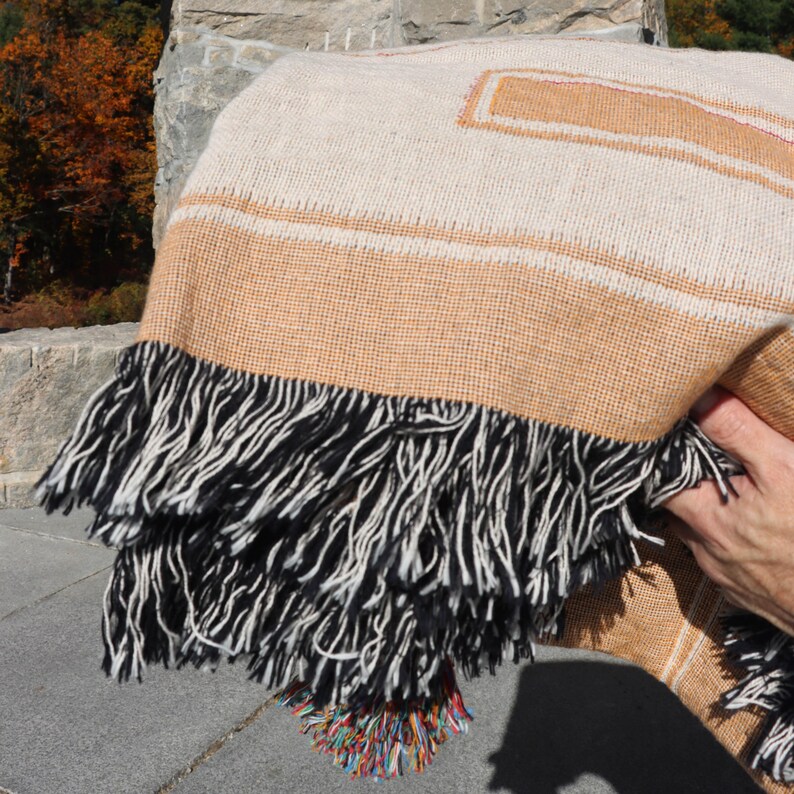 May include: A woven blanket with a cream-colored background and tan stripes and accents. The blanket has a thick fringe of black and white threads along the bottom edge. A small section of colorful threads is visible at the bottom. The blanket is folded.