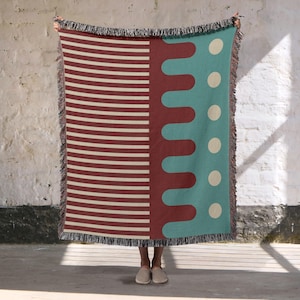 May include: A woven throw blanket with a geometric design. The blanket features a section of horizontal cream stripes, a section of maroon wavy lines, and a section of teal with cream-coloured circles. The blanket has a fringed edge.