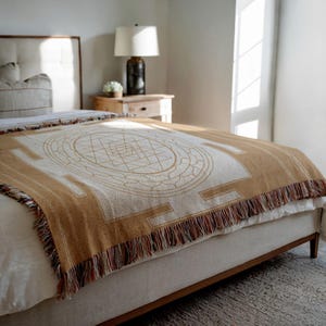 May include: A tan and cream-colored throw blanket with a geometric design and fringe detail. The blanket is draped over a bed, showcasing its intricate pattern and texture. The design features a central circular motif and rectangular borders.