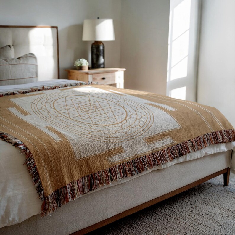 May include: A tan and cream-colored throw blanket with a geometric design and fringe detail. The blanket is draped over a bed, showcasing its intricate pattern and texture. The design features a central circular motif and rectangular borders.