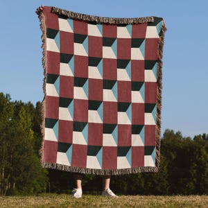 Peut inclure: Une couverture à motifs géométriques avec un motif de cubes 3D répétés en bordeaux, noir, blanc et bleu clair. La couverture a un bord à franges et est tenue contre un fond de ciel bleu.