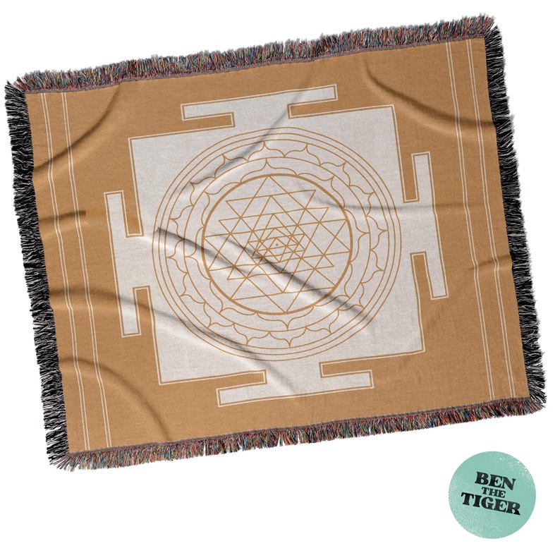 May include: A tan and cream-colored woven throw blanket with a geometric design. The blanket features a central geometric pattern in cream, surrounded by a tan border and fringe. The design includes a series of triangles and lines.