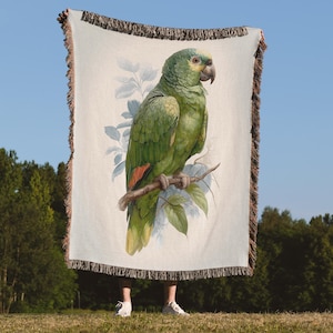 May include: A cream-colored throw blanket featuring a detailed illustration of a green parrot perched on a branch. The parrot has yellow and red accents. The blanket has a fringed border and is held up against a blue sky.